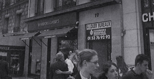 A black-and-white photograph of a busy, walkable street in Paris with pedestrians and shops visible.