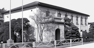 Black-and-white image of a classic building, the Odawara Cosmetics Factory, surrounded by well-trimmed shrubbery and trees.
