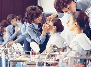 Many women, standing in a row in blue uniforms apply makeup to customers sitting in chairs facing tables with mirrors and supplies on top.