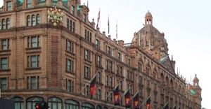 A large, brown, classic building, Harrods department store in London, adorned with a wide variety of flags, viewed from the street level.