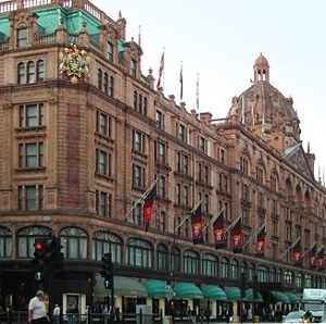 A large, brown, classic building, Harrods department store in London, adorned with a wide variety of flags, viewed from the street level.