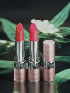 Three lipsticks standing on a table, one closed, two open revealing shades of red. A green and white plant appears in the background.