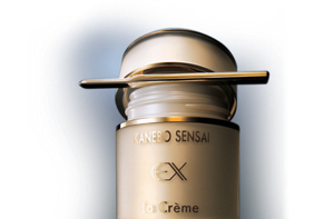 A beige and gold cosmetics bottle with the lid half-opened and text that reads, “KANEBO SENSAI EX” and “La Crème”.