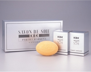 Three silver-colored packages with a bar of soap in front from the SAVON DE SOIE silk soap product line launched in the 1930s.