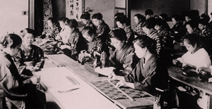Many women in a classroom participating educational activities with sewing machines, textiles, and fabrics appearing on the tables.