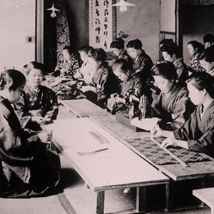 Many women in a classroom participating educational activities with sewing machines, textiles, and fabrics appearing on the tables.