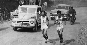 Two runners in a black-and-white photo competing in a race on a gravel road followed by three cars and a motorcycle.