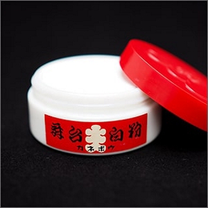 Round, white, and red Kabuki foundation case. On its side rests the open lid revealing the white makeup contained inside.