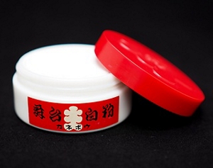 Round, white, and red Kabuki foundation case. On its side rests the open lid revealing the white makeup contained inside.