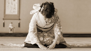 A sepia image of a Japanese woman in traditional Japanese clothing bowing in a traditional Japanese room.