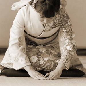 A sepia image of a Japanese woman in traditional Japanese clothing bowing in a traditional Japanese room.