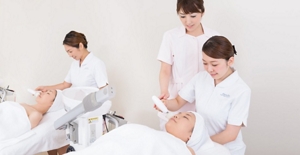 Three women in white uniforms providing beauty services to two women in white towels laying on separate tables.