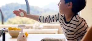 A young boy at a sink with foaming hand soap on his hand near a bottle of My Kirei by KAO soap.