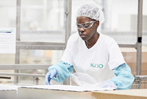 A woman working on an assembly line. She is wearing safety glasses and gloves with a white, KAO-branded smock.