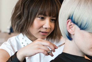 A young hair stylist trimming bleach blond hair with dark blue edges.