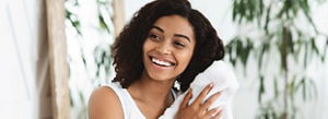 A young black woman with a fresh face and bright smile. She is wearing a white tank top, towel-drying her hair in a tranquil setting.