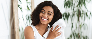 A young black woman with a fresh face and bright smile. She is wearing a white tank top, towel-drying her hair in a tranquil setting.