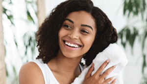 A young black woman with a fresh face and bright smile. She is wearing a white tank top, towel-drying her hair in a tranquil setting.