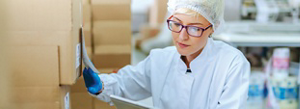 A middle-aged Caucasian woman wearing a hair net, latex gloves and white jacket comparing a label on a box to something she is holding.