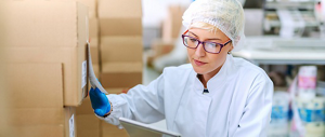 A middle-aged Caucasian woman wearing a hair net, latex gloves and white jacket comparing a label on a box to something she is holding.
