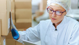 A middle-aged Caucasian woman wearing a hair net, latex gloves and white jacket comparing a label on a box to something she is holding.