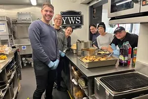 Employees in the kitchen prepare meals and place them on the counter for young people to gather around.