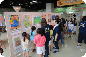 こども達の作品を展示する展示会場の写真。絵の周りには大人とこども達が集まっています。