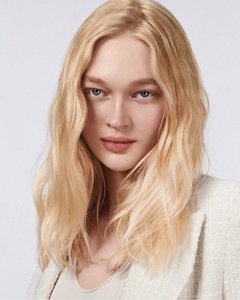 a blonde white woman with long hair is looking straight into the camera while holding a goldwell stylesign smooth product in her hands on a white background