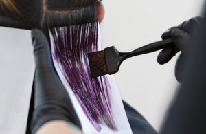 A hairdresser performing the goldwell elumen creative looks & freehand technique service by dying a persons long black hair purple.