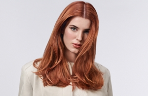 A woman with straight nouveau copper-colored hair, dyed with Goldwell Topchic, standing in front of a grey background.
