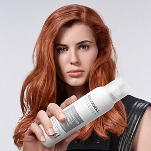 a red haired european woman with a black dress looking straight in the camera, while holding a goldwell stylesign hairspray on a white background