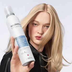 a blonde white woman with long hair is looking straight into the camera while holding a goldwell stylesign volume product in her hands on a white background