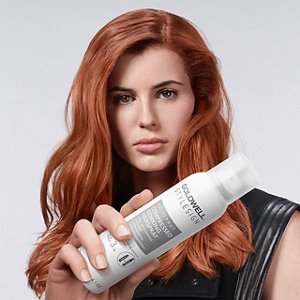 a red haired european woman with a black dress looking straight in the camera, while holding a goldwell stylesign hairspray on a white background