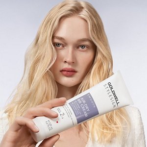 a blonde white woman with long hair is looking straight into the camera while holding a goldwell stylesign smooth product in her hands on a white background