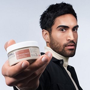 a black haired man with a beard and a black jacket is looking straight into the camera while holding a goldwell stylesign texture product in his hands on a white background