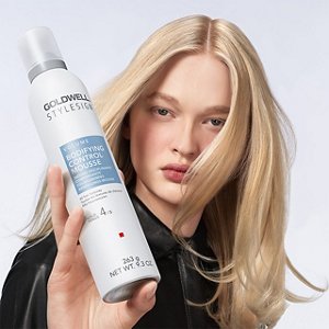 a blonde white woman with long hair is looking straight into the camera while holding a goldwell stylesign volume product in her hands on a white background