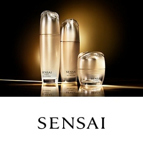 Three Sensai products of various sizes in beige packaging positioned side-by-side surrounded by a white fan in the background. 