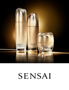 Three Sensai products of various sizes in beige packaging positioned side-by-side surrounded by a white fan in the background. 