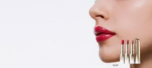 Quattro rossetti di diverse tonalità sono disposti in supporti dorati davanti a un primo piano di labbra rosse e lucide