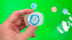化石が描かれたペットボトルのキャップが入っている氷を持っている手。背景は緑のお湯の画像