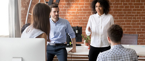 A young black woman wearing a white blouse and black pants is speaking to co-workers in a contemporary office space.