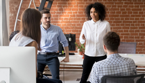 A young black woman wearing a white blouse and black pants is speaking to co-workers in a contemporary office space.