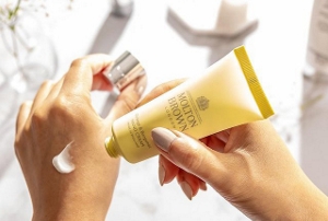 A close-up of well-manicured hands applying lotion from a yellow tube with Molton Brown logo.