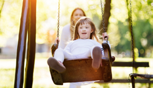 A young girl with Down syndrome being pushed on a swing by an out-of-focus person in the background.