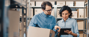 A man and a woman in a warehouse surrounded by boxes. The man is pointing to a tablet while they discuss what is on screen.