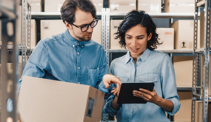 A man and a woman in a warehouse surrounded by boxes. The man is pointing to a tablet while they discuss what is on screen.