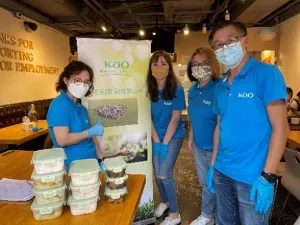 4名義工站在印有花王標誌的直立展示架旁合照，旁邊的桌上擺放著飯盒。