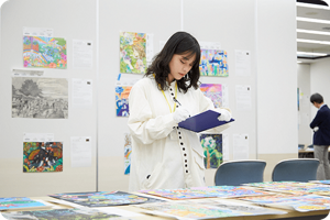 白い手袋をつけた審査員が机の上に並べられたこどもたちの作品を丁寧に審査している様子