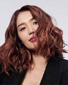 a red haired asian woman with a black blazer is holding a goldwell stylesign heat styling product, while looking in the camera in front of a white background