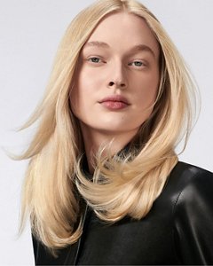 a blonde white woman with long hair is looking straight into the camera while holding a goldwell stylesign volume product in her hands on a white background